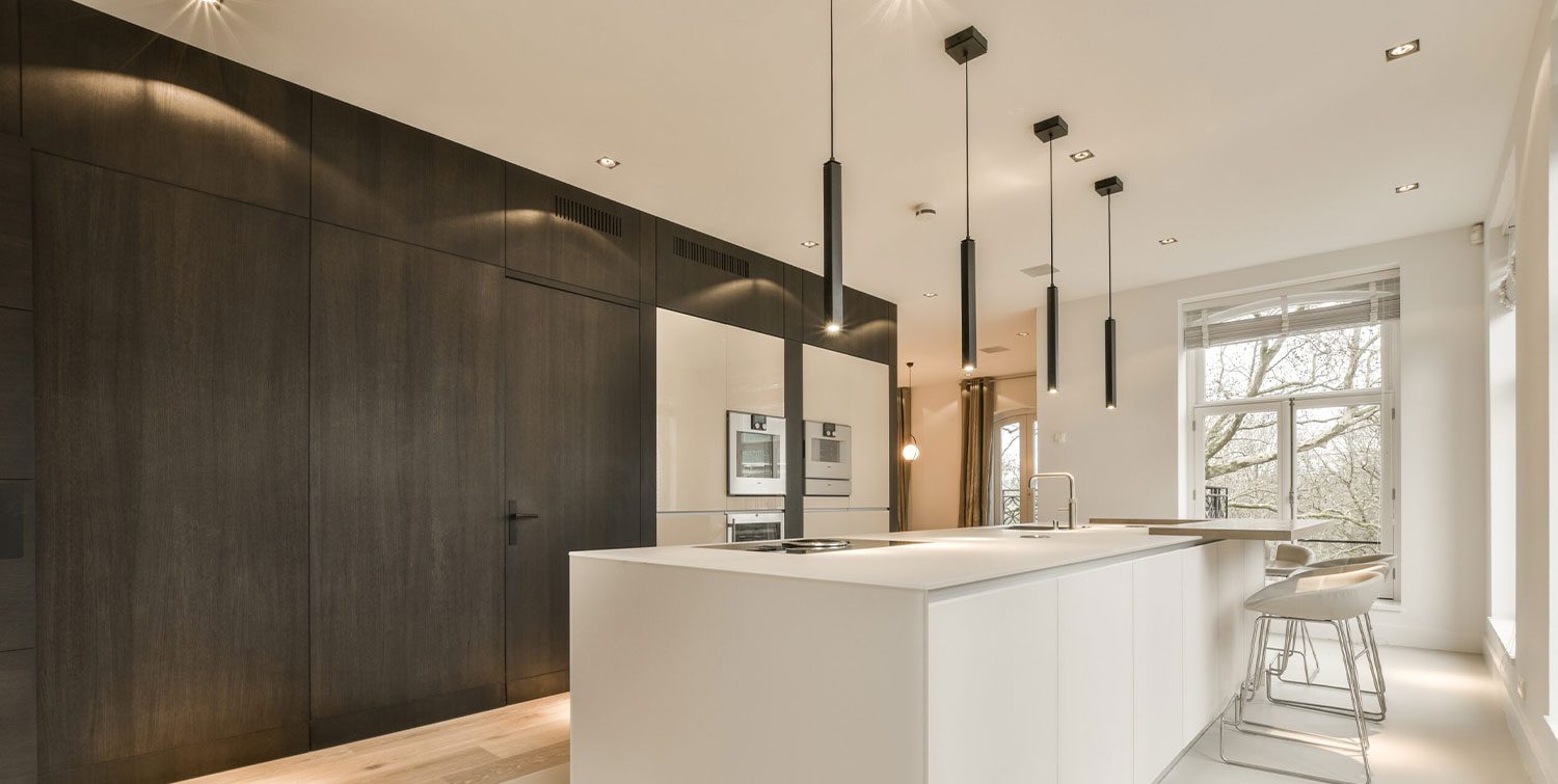 stock-photo-a-modern-kitchen-with-white-counters-and-dark-wood-cabinets-in-the-room-is-lit-by-recessed-light-2292055671-1 A modern kitchen features a large white island with barstools, built-in ovens, and sleek dark cabinetry. Pendant lights hang from the ceiling providing illumination over the countertops.